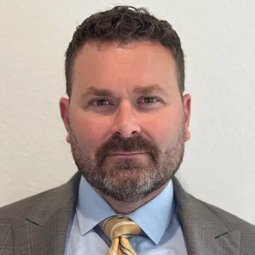 Headshot of Jared Stephenson, VP of Roofing Systems at NanoTech Materials — a man with short dark hair and a salt-and-pepper beard, wearing a gray plaid suit jacket, light blue shirt, and gold tie against a plain cream background